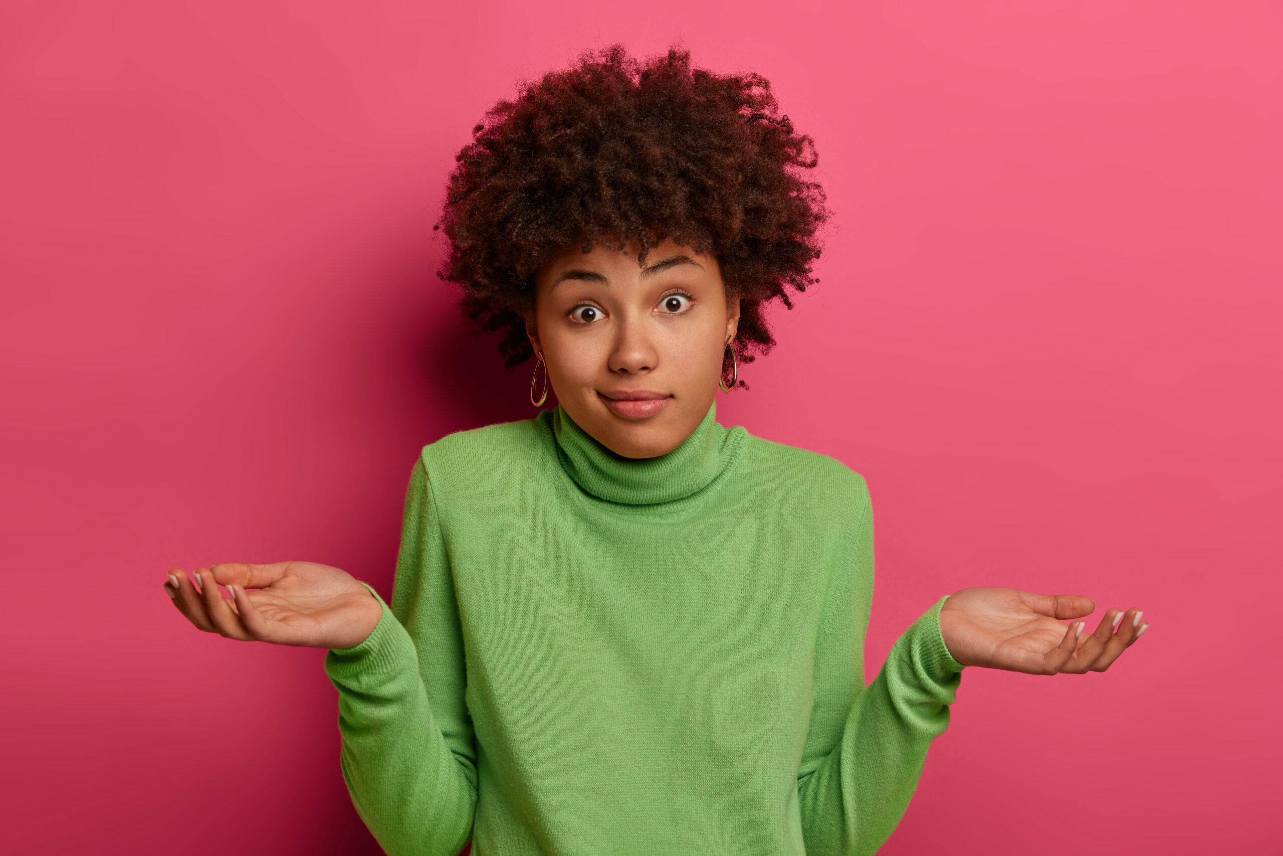 Indifferent confused black woman spreads palms confusingly, has doubts, wears green turtleneck, has hesitant face expression, isolated over pink background, has no idea, puzzled to give answer